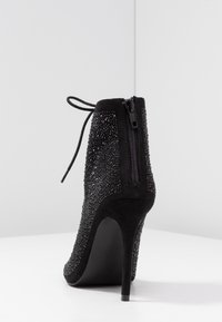 Zwarte enkellaars met hoge naaldhak, suède materiaal en versierd met fonkelende strass-steentjes. Achterritssluiting en veterdetail.