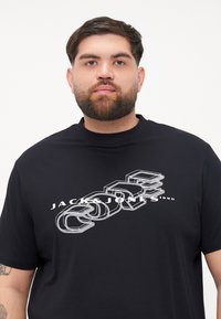 Juodas marškinėlis su grafiniu dizainu, kuriame yra trimatiai raidžių elementai ir prekės ženklas "JACK & JONES" balta spalva, trumpomis rankovėmis.