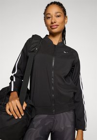 Black zip-up jacket with white side stripes, made from lightweight material. Worn over patterned leggings, paired with a black duffel bag.