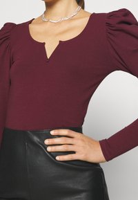 Burgundy pitkähihainen paita syvässä V-mallissa ja kerroksellisissa puffihihnoissa, yhdistettynä korkeavyötäröiseen mustaan tekonahkahameeseen.