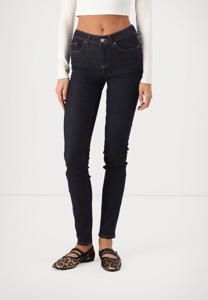 Jeans de tiro alto azul oscuro con cinco bolsillos, costuras naranjas, ajuste slim y combinados con unos zapatos planos de estampado animal marrón con tiras negras.