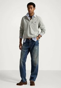 Jovem de pé com uma mão no bolso, vestindo uma camisa polo cinza de manga longa sobre uma camisa riscada azul, jeans azuis e sapatos castanhos.