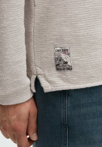 Helles graues, strukturiertes Sweatshirt mit einem Camp David Label, seitlichen Schlitzen und gerippten Bündchen, kombiniert mit dunkelblauen Jeans.