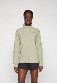 Light green jacket with a high collar, featuring orange and black logos. It has a zip front and hand pockets, made from a lightweight material.