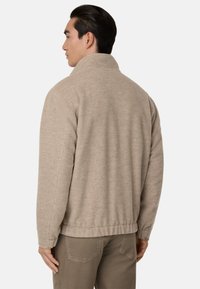 Beige flísová bunda s vysokým límcem, žebrovaným lemem a dlouhými rukávy. Měkká textura, volný střih a minimální prošívání na zadní straně.
