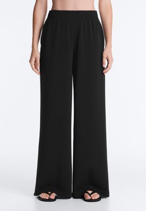 Personne portant un pantalon large noir avec une taille élastique, debout pieds nus sur un fond blanc.