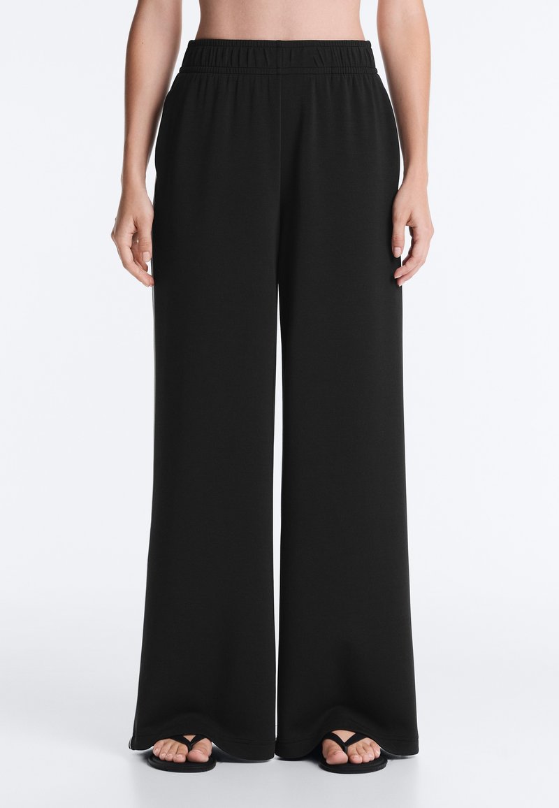 Personne portant un pantalon large noir avec une taille élastique, debout pieds nus sur un fond blanc.