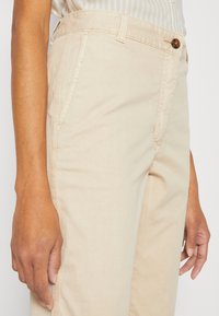 Beżowe bawełniane spodnie z przednimi kieszeniami, zapięciem na guziki i detalami przeszyć. Gładka faktura i prosty krój nogawek.
