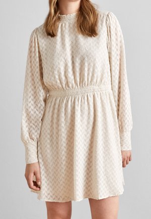 Vrouw draagt een crèmekleurige, getextureerde jurk met lange mouwen, elastische taille en manchetdetails, staande tegen een effen lichtgrijze achtergrond.