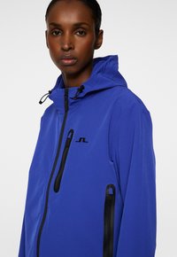 Giacca blu con cappuccio, realizzata in tessuto liscio. Presenta zip nere e un logo sul petto. Due tasche laterali con zip.