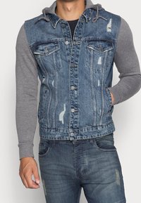 Denim vest med grå lange ærmer, slidt struktur, knaplukning og to brystlommer. Hætte med snøringer fastgjort ved halslinjen.