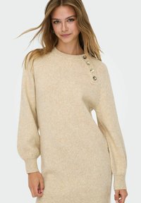 Beige gebreide jurk met ronde hals, lange pofmouwen en vijf decoratieve gouden knopen op de schouder. Geribbelde manchetten en zoom.