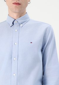 Lichtblauwe button-up shirt met een kraag, voorzien van witte knopen en een klein geborduurd logo in rood, wit en blauw op de borst.
