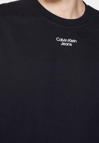 Sudadera negra de cuello redondo con el pequeño logotipo blanco "Calvin Klein Jeans" centrado debajo del escote en el pecho.