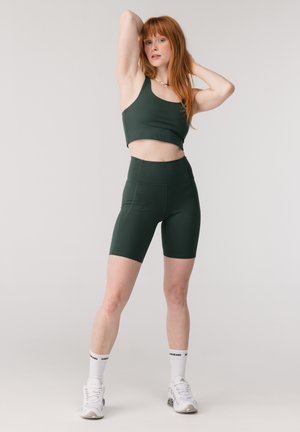 Žena s červenými vlasmi v tmavozelenom krátkom topu a cyklistických šortkách, biele ponožky a biele tenisky, pózuje s rukami zdvihnutými a ohnutými.