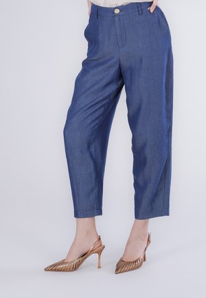 Person iført blå bukser med høj talje og tilspidset ben, der har knap og lynlås foran, kombineret med metalliske slingback-hæle i rosaguld med spids tå.