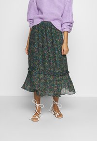 Korkea kukallinen midihame, jossa on kerroksellinen röyhelöhelma, tummalla taustalla värikkäitä kukkakuvioita, käytetään lilassa neulepaidassa ja valkoisissa hihna-sandaaleissa.