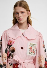 Νέα γυναίκα που φοράει ένα ροζ φλοράλ καμπαρντίνα με κυματιστό γιακά, δεμένη ζώνη και ένα έμβλημα "Barbour Farm Rio" στο στήθος.