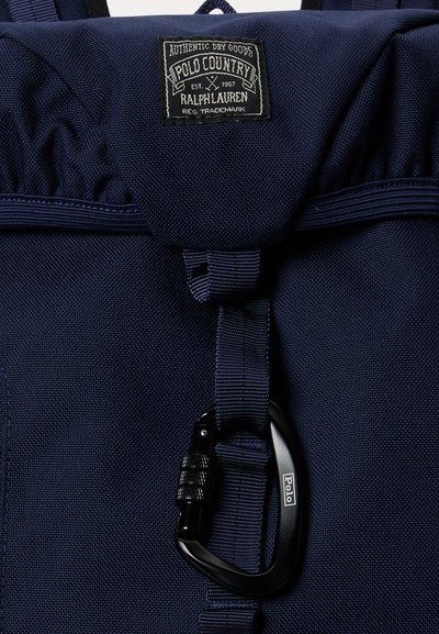 Σακίδιο Polo Country σε ναυτικό μπλε χρώμα με μπροστινό καπάκι, ραμμένο λογότυπο σε μορφή μπάλωμα και μαύρο κλειδωμένο καραμπίνερ προσαρτημένο σε κάθετο ιμάντα.