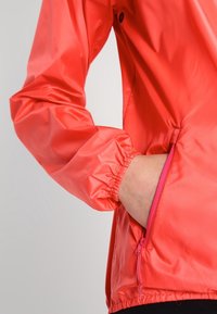 Veste légère rouge avec des manches élastiques, poche zippée sur le côté et une texture lisse et brillante. Présente un accent rose sur la fermeture éclair.