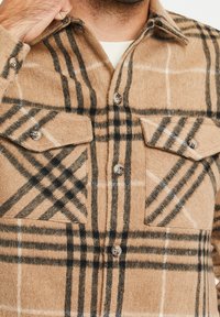 Chemise en flanelle à carreaux marron avec des rayures beiges et noires, deux poches avant, et des fermetures à boutons sur un tissu doux et texturé.