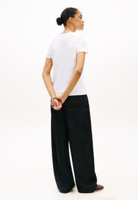 Kobieta z włosami związanymi do tyłu, ubrana w białą koszulę z krótkim rękawem, ciemne szerokie dżinsy i brązowe buty, stoi z rękami splecionymi za plecami.