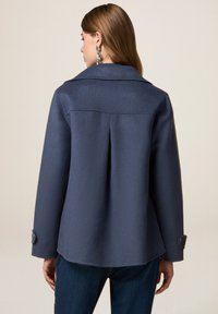 Giacca in lana blu navy con colletto classico, piega sul retro e polsini con bottone. Tessuto liscio con un design pulito e sartoriale.