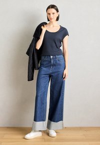Laine navy ribbed lyhyet hihat -paita yhdistettynä leveälahkeisiin denim-farkkuihin, joissa on purjeraita ja harmaita yksityiskohtia. Henkilö pitää mustaa takkia.
