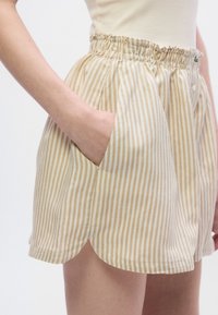 Shorts beiges à rayures avec une taille élastique et des poches latérales ; le tissu est léger et légèrement texturé, avec un ourlet incurvé.