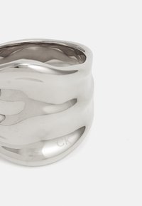 Anello in metallo argentato scolpito con una texture fluida e ondulata; presenta una finitura lucida e il logo "CK" inciso all'interno della banda.