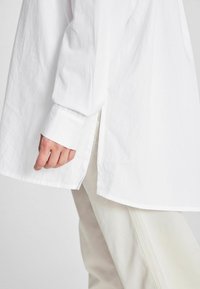 Camicia bianca a maniche lunghe realizzata in tessuto morbido, con orlo arrotondato, spacchi laterali e polsino abbottonato. Abbinata a pantaloni chiari.