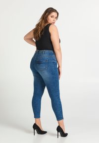 Jeans de denim azul de tiro alto con un ajuste slim, costuras detalladas y bolsillos traseros, combinados con un top sin mangas negro y tacones altos negros.