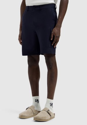 Man wearing navy cargo shorts, white socks with black lettering, and beige slip-on sandals against a plain background.