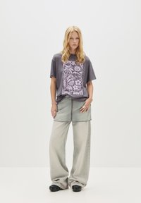Grafisk grå t-shirt med et "Tropical Lines" print i pink, parret med brede lysegrå denimbukser og sort fodtøj.