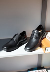 Paar schwarze Lederschuhe mit Schnürsenkeln, die auf einem weißen Regal stehen, ein Schuh liegt auf einem orangefarbenen Buch.