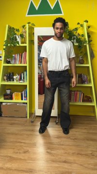 Jeans de mezclilla negra con corte de pierna recta, usados con una camiseta blanca lisa y zapatos negros. El fondo presenta estantes verdes y un espejo.