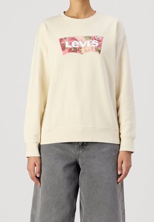Persoon draagt een crèmekleurige Levi's trui met een roze bloemenlogo en losse grijze spijkerbroek, staand tegen een effen achtergrond.