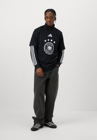 Camisa desportiva preta com colarinho alto, emblema da seleção alemã de futebol em branco e cinco estrelas; apresenta riscas brancas da Adidas nos braços; combinada com jeans escuros.