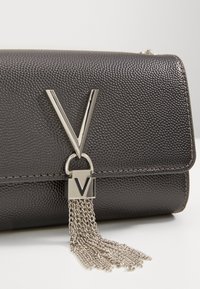 Clutch in pelle nera con texture, dotata di chiusura a forma di V in argento e dettaglio con nappe a catena, su una superficie bianca.