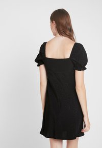 Musta lyhyt mekko neliönmuotoisella selkämyksellä, teksturoitu kangas, puffihihat ja levenevä helma. Kankaassa on hienovarainen kuvio.