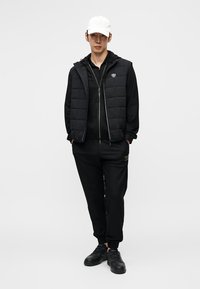 Juodas paminkštintas liemenė dėvima ant juodos striukės su užtrauktuku, derinama su juodomis sportinėmis kelnėmis ir juodais sportbačiais. Baltas beisbolo kepuraitė ir subtilūs prekės ženklo detalės.