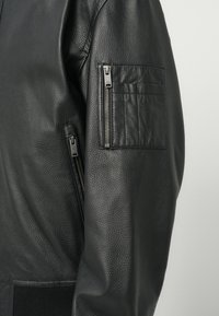 Melns teksturēts ādas jaka piedurknes ar vertikālu sudraba rāvējslēdzēja kabatu un mazu taisnstūra formālu rāvējslēdzēja nodalījumu augšējā rokā.