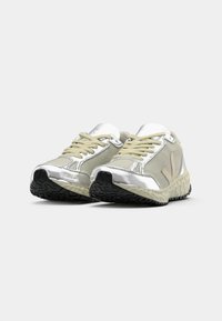 Sportowe buty do biegania z beżową siateczką, metalicznie srebrnymi akcentami, beżowymi sznurowadłami i teksturowaną czarną gumową podeszwą.