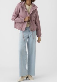 Kvinna iklädd en dammig rosa huvtröja över en beige tröja, ljusblå vida jeans och krämfärgade ankelstövlar, stående mot en vit bakgrund.