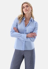 Light blue button-up shirt with vertical white stripes, featuring a chest pocket and structured collar, paired with gray textured trousers.