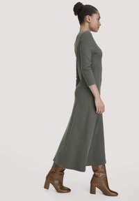 Langes, enganliegendes olivgrünes Kleid mit dreiviertellangen Ärmeln, gerippter Struktur und fließendem Saum, kombiniert mit braunen Stiefeln mit Absatz.