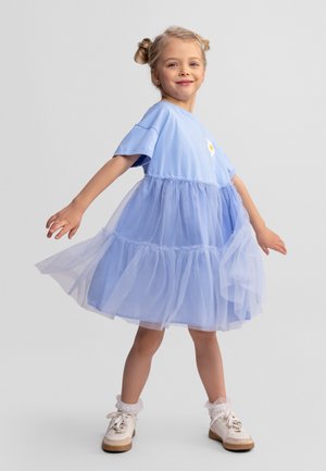 Young girl spinning in a knee-length light blue dress with tulle layers, wearing white sneakers and smiling against a plain background.