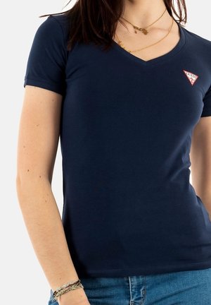 Frau trägt dunkelblaues V-Ausschnitt-T-Shirt mit kleinem roten und weißen Guess-Logo auf der Brust, kombiniert mit blauer Jeans und geschichteten goldenen Halsketten.