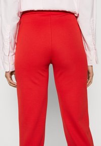 Pantalon rouge extensible avec une taille haute, présentant une texture lisse et une coupe ajustée. La vue de dos montre des coutures minimales visibles et aucune poche.