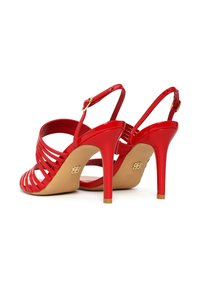 Sandalias rojas de tacón alto con múltiples tiras, hebillas en el tobillo y suelas beige, mostradas desde un ángulo trasero sobre un fondo blanco.